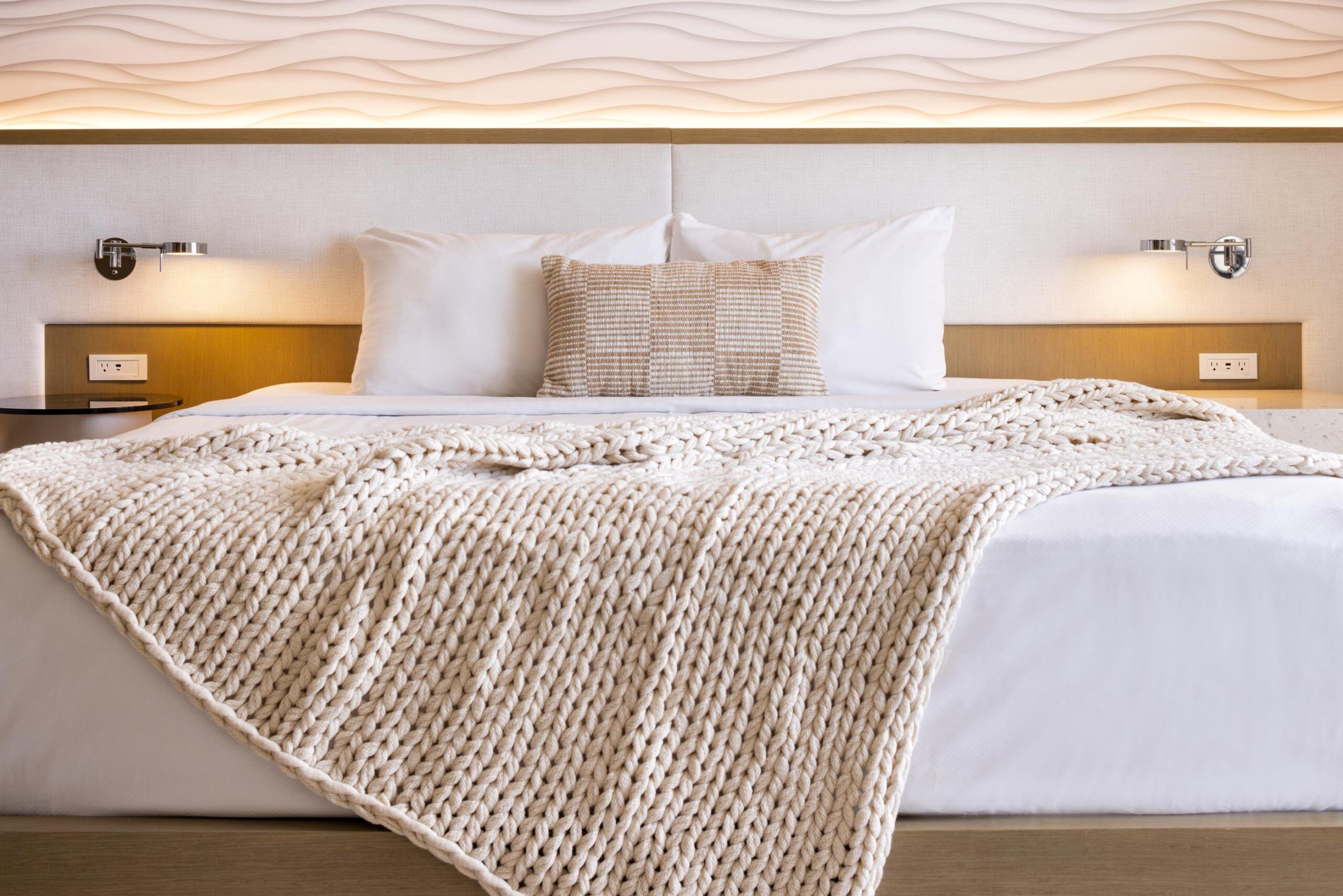 A neatly made standard bed with white sheets, a beige knit throw blanket, a textured pillow, and wall-mounted reading lights.