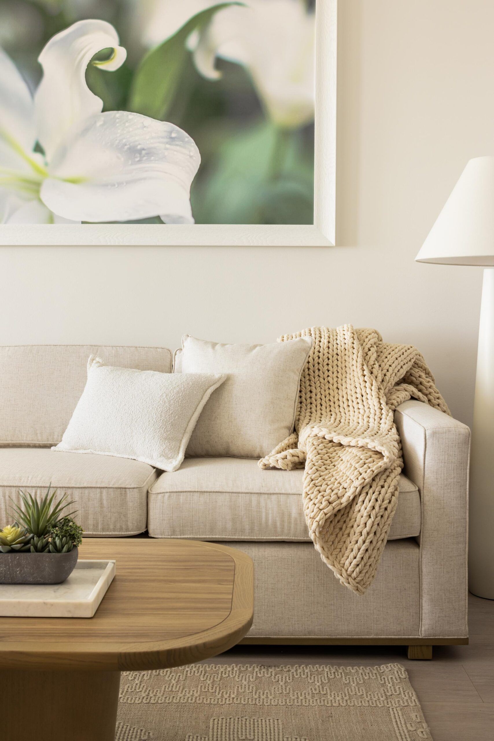 A cozy living room with a beige sofa, cushions, a knitted blanket, a wooden table with a plant, and floral wall art offers direct access to the balcony for those enjoying an Intracoastal Queen view.