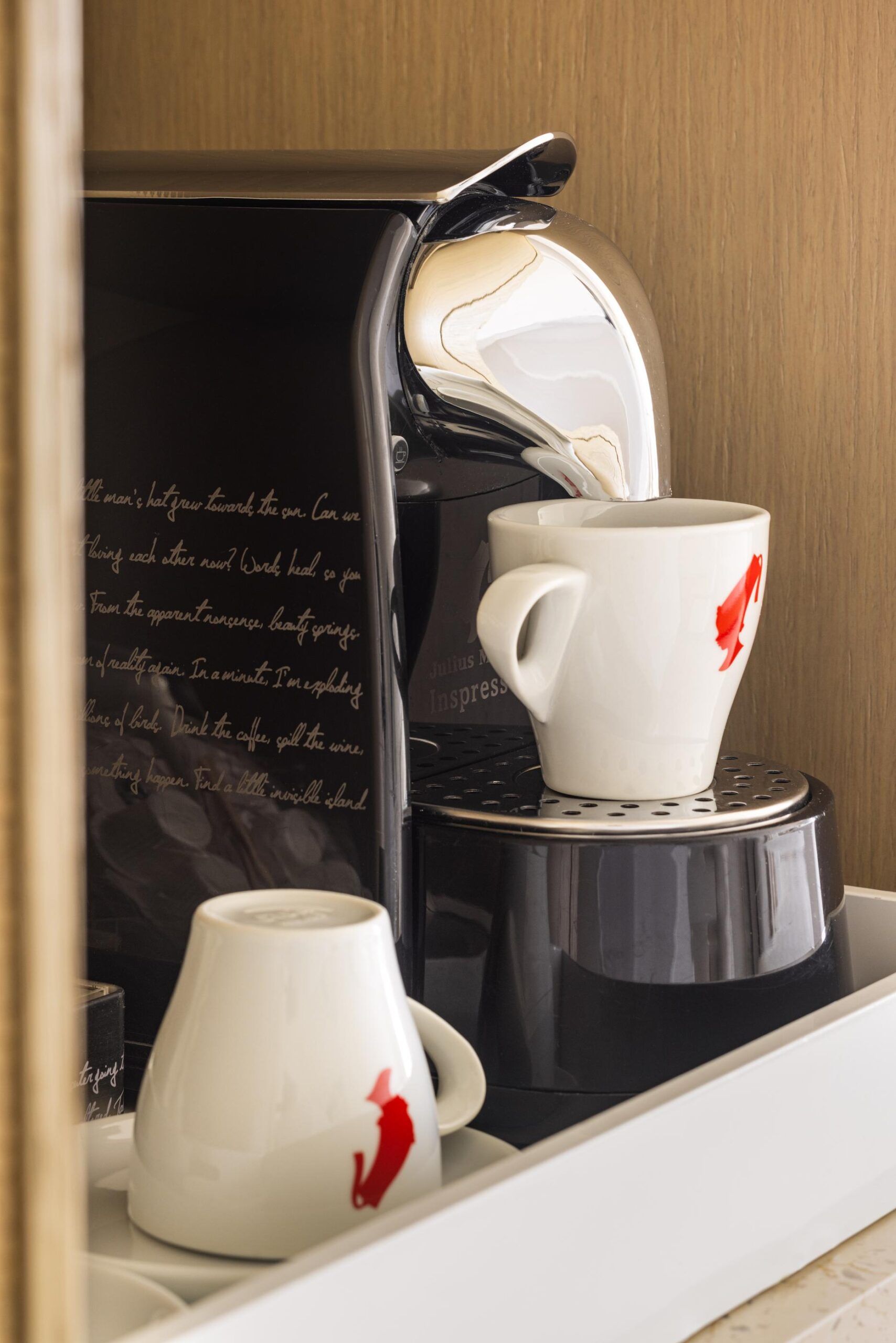 Black pod coffee machine with a white coffee cup and an overturned cup, both featuring red fish illustrations, set on a tray—all perfectly suited for your Intracoastal Queen balcony mornings.