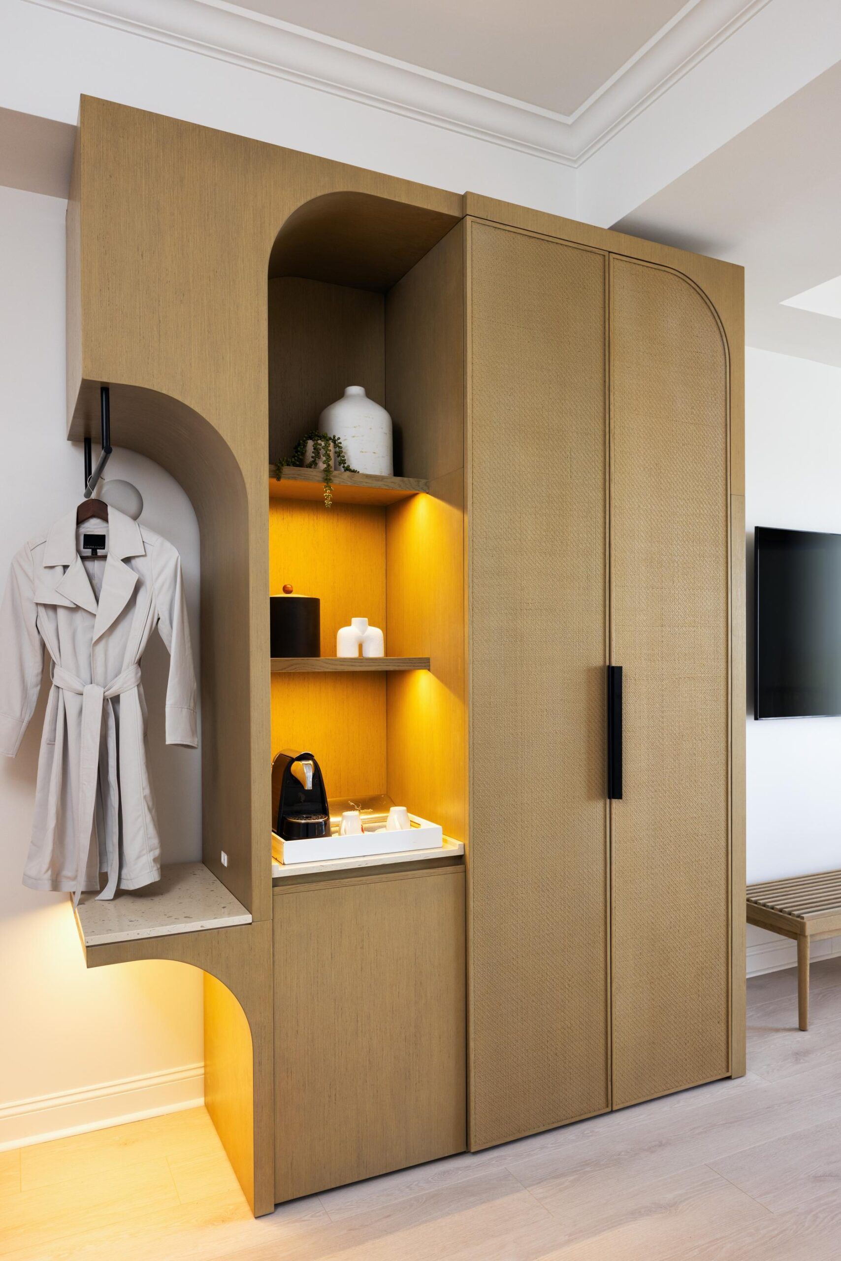 Modern wooden wardrobe with an illuminated built-in shelf featuring a coffee maker, cups, decor items, and a hanging white robe fit for a Queen Standard.