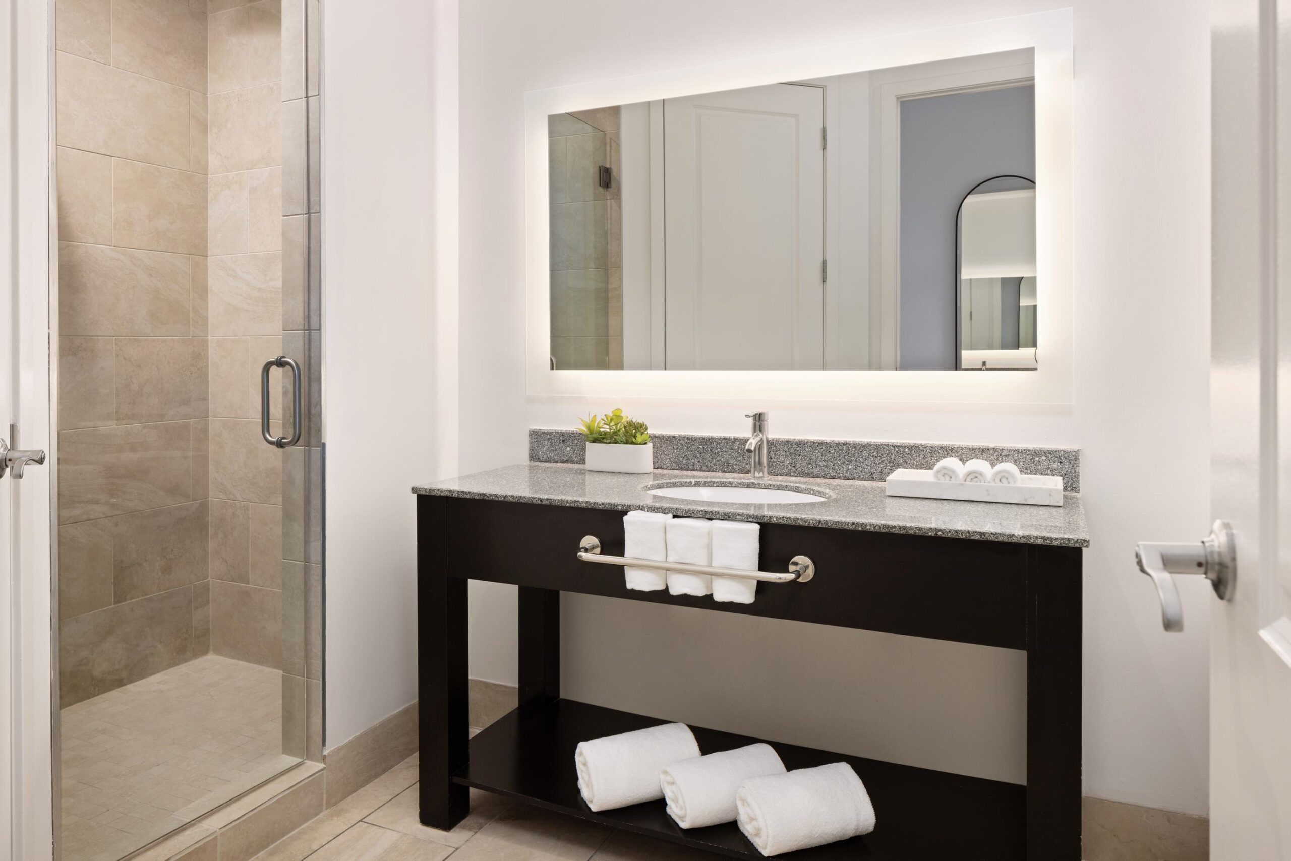 Modern bathroom with a glass shower, large backlit mirror, sink on a dark vanity, and white towels on shelves. This stylish space is part of the luxurious Queen Room that offers a breathtaking view from its private balcony.