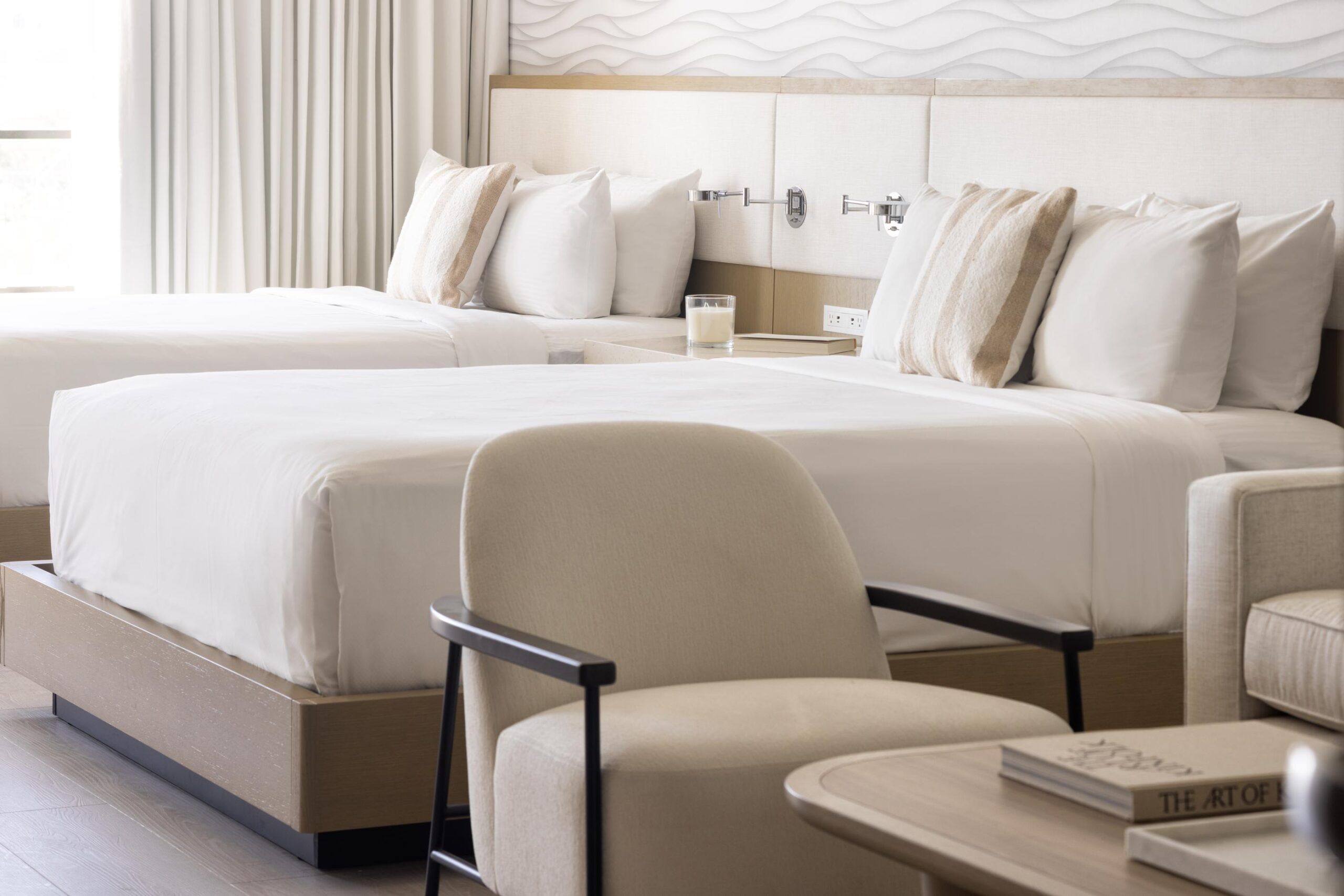 Bright Queen Standard hotel room with two neatly made double beds, a lamp, a beige armchair, and a small round table with a book.