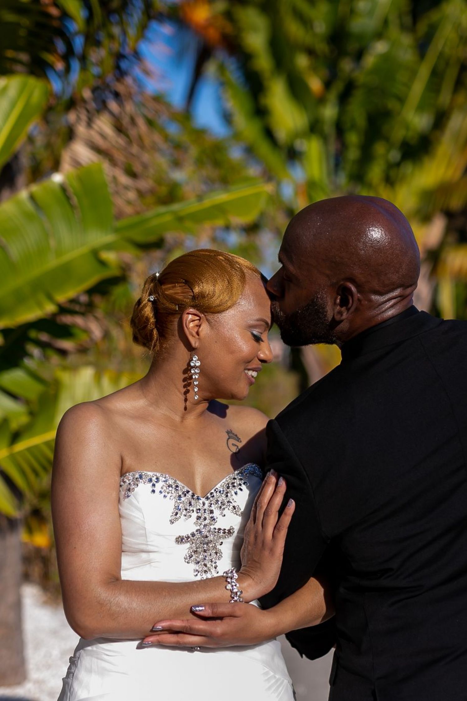 Dulce pareja vestida de novia compartiendo un beso al aire libre con plantas tropicales detrás